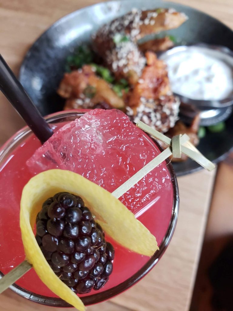 alt text = "pink cocktail on the rocks with black berry and lemon for garnish, chicken wings in the background"