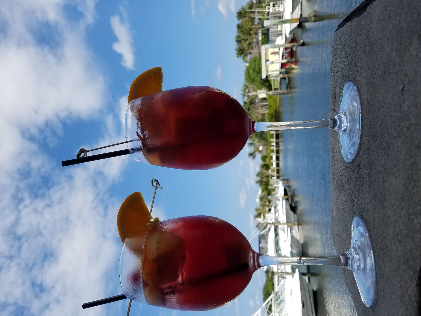 alt text = "two refreshing long stemmed glasses of red sangria garnished with fruit with a marina in the background