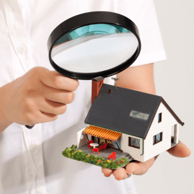 alt="Woman in a white blowse holding a magnifying glass over a wooden model house to symbolize a home inspection."